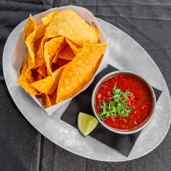 Crispy tortilla chips in a paper-lined basket next to a bowl of salsa and a lime wedge.