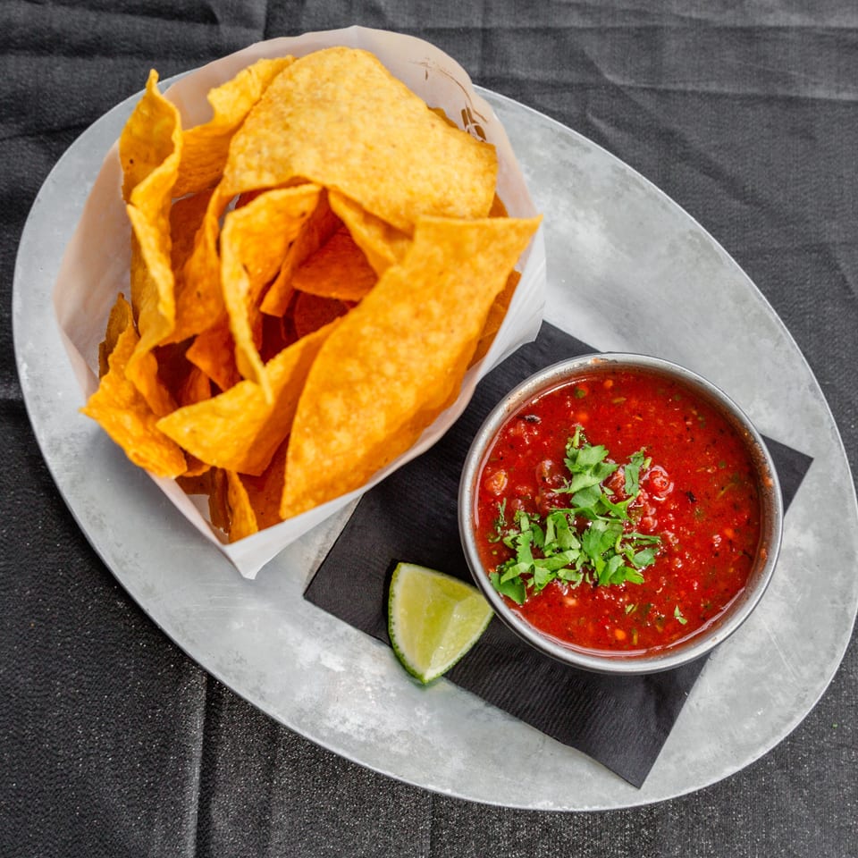 Crispy tortilla chips in a paper-lined basket next to a bowl of salsa and a lime wedge.