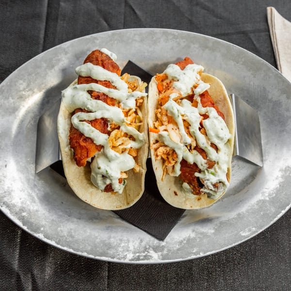 Two Vida Taco Bar fish tacos with slaw and creamy sauce on a metal plate.