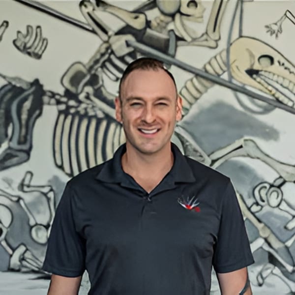 Smiling man in a dark polo shirt with a logo, standing before a mural of skeletons.