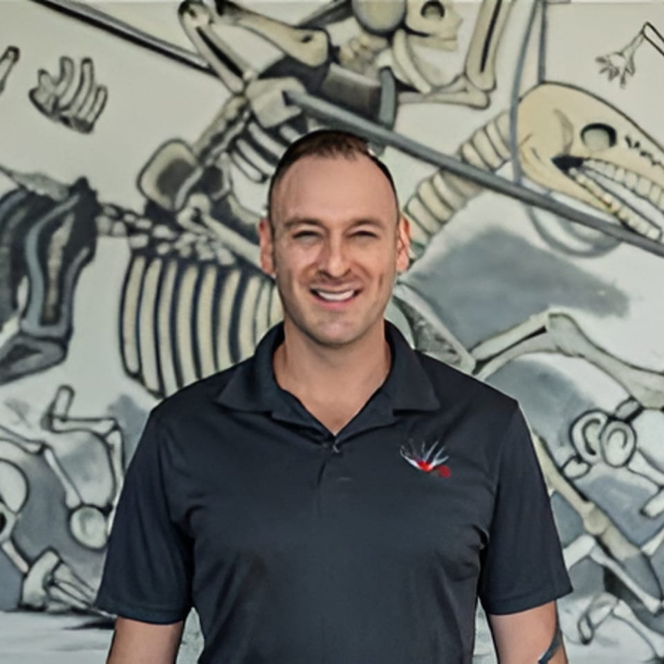 Smiling man in a dark polo shirt with a logo, standing before a mural of skeletons.