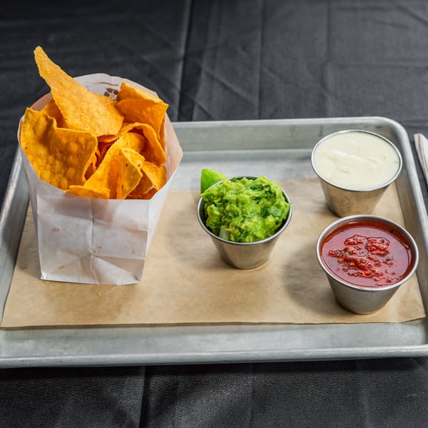 A tray of tortilla chips in a paper bag with sides of guacamole, salsa, and queso.