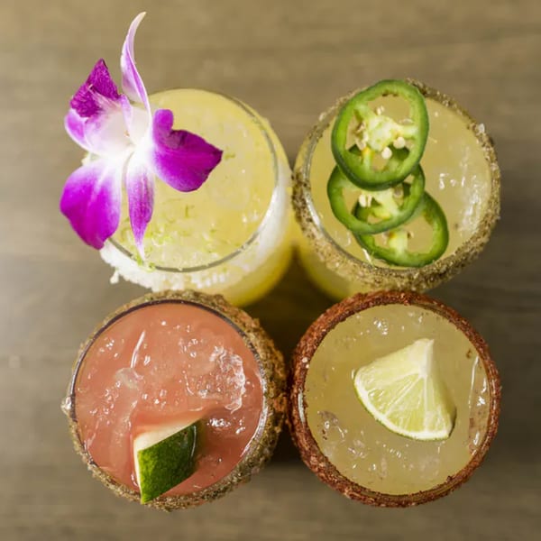 Four Vida Taco Bar margaritas, two yellow, one pink, and one light yellow, garnished with fruit and spices.