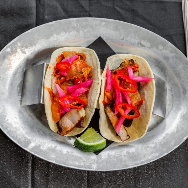 Two Vida Tacos with pork belly, kimchi, pickled onions, and red chilis on a metal plate.